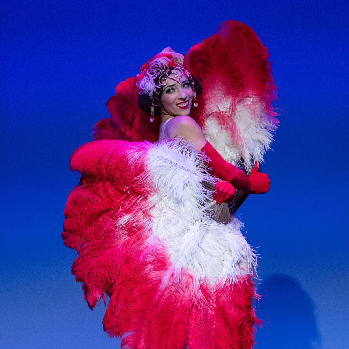 Award-winning and internationally touring burlesque artist and showgirl, Chanelle du Mai from Paris, France performing with Adèle Wolf Productions in Oklahoma City. She is posed with red and white ostrich feather fans and wearing a rhinestoned showgirl headdress.