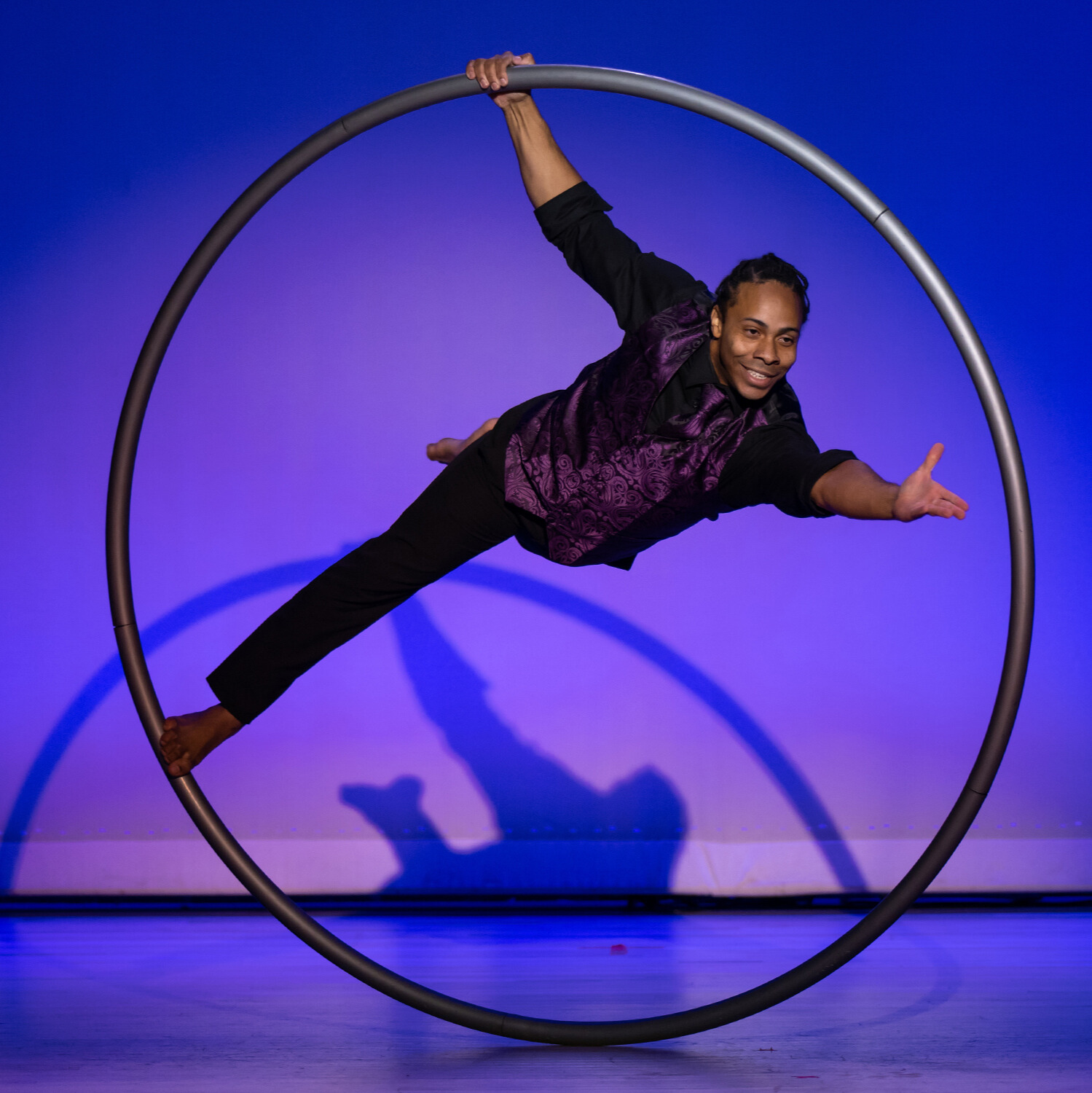 Renowned cyr wheel artist, Nick Hodge, from Las Vegas performing with Adèle Wolf Productions in Oklahoma City. Nick is wearing black slacks with a black button up shirt and purple vest. He poses with his left leg pointed behind him and his left arm extended to the crowd.