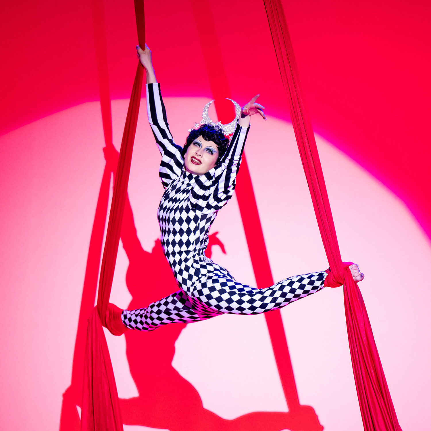 Award-winning aerialist and local resident artist, Olympe, performing with Adèle Wolf Productions in Oklahoma City. They are wearing a harlequin style black and white bodysuit and rhinestoned moon headdress while posed in the splits on red aerial silks.